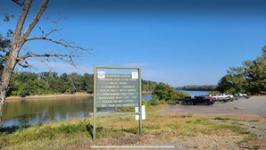 A sign by a lake with a parking area, trees, and a clear sky. - Riverside Lodge (Clarendon)