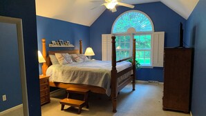 Un dormitorio con cama con dosel, un taburete de madera, una mesita de noche con lámpara, una ventana con contraventanas y un ventilador de techo.