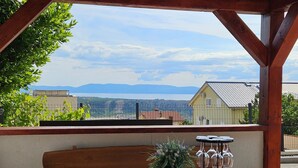 A wooden table with a bowl of fruit, a potted plant, and wine glasses on a balcony overlooking a scenic view of mountains and houses.