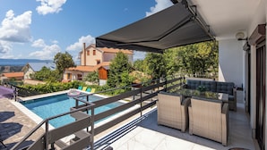 A rooftop pool area with a view of houses and trees, a covered seating area, and a poolside table.