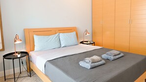 A neatly made bed with a grey blanket and two pillows, a small round table with a lamp, and a large wardrobe.