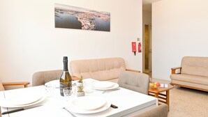 A dining area with a table set for two, a bottle of wine, and a small plant. There is a sofa and a chair in the background.