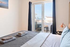 Un cuarto de hotel con cama, una mesita, una silla y vista al mar a través de una puerta corrediza de vidrio.