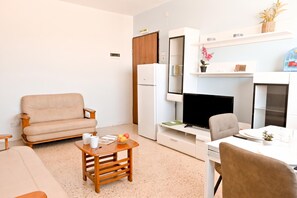 A compact living space with a sofa, a small wooden table with a bowl of fruit, a television, and a dining area with a chair and table.