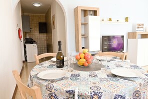 Un coin repas avec une table dressée pour deux personnes, un saladier de fruits, une bouteille d’huile d’olive et un téléviseur en arrière-plan.