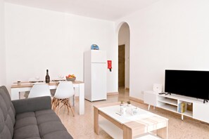 A modern living room with a sofa, a dining table with chairs, a kitchen area with a refrigerator, and a television on a stand.