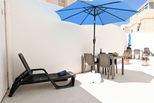 A patio with a table, chairs, a blue umbrella, and a lounge chair.