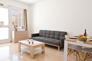 A modern living room with a grey sofa, a wooden coffee table, a dining table with chairs, and a small plant on the windowsill.