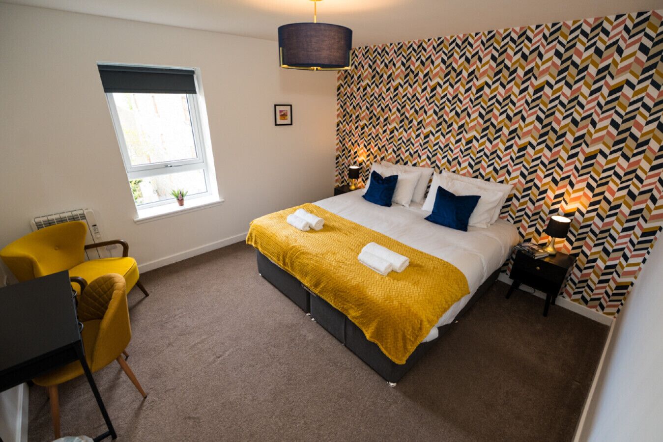 Une chambre d’hôtel avec un lit, un bureau, une chaise, une fenêtre et un mur décoré de motifs.