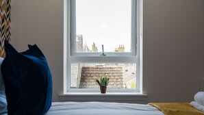 Un dormitorio con una cama, una ventana que da a vista de edificios, una planta en maceta en el alféizar y una bolsa azul sobre la cama.