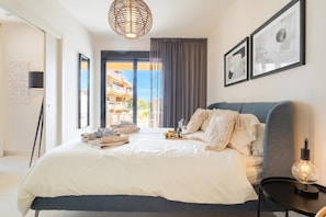A modern bedroom with a large bed, a bedside table with a lamp, and a view of the outside.