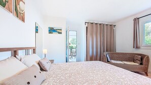Ein Schlafzimmer mit einem großen Bett, einem Korbssessel, einem Schreibtisch und einem Fenster mit Vorhängen.