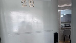 Uma porta com o número 2B, maçaneta moderna e, ao fundo, um banheiro visível com espelho e pia.
