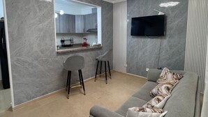 Uma sala de estar moderna com um sofá cinzento, um espaço de jantar com bancada de cozinha e uma TV de tela plana na parede.