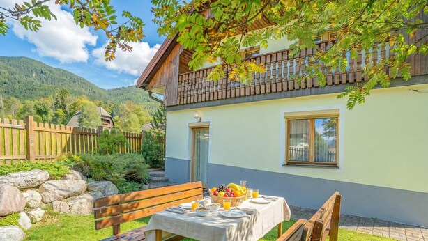 A two-story chalet with a balcony, a dining table set with breakfast, and a stone wall in the background.