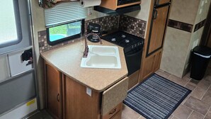 A compact RV kitchen with wooden cabinets, a microwave, a sink, and a stove. - RV Bunkhouse on the River (Mason City)