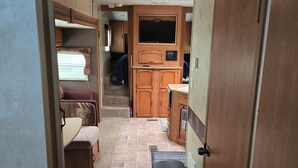 A compact RV interior with a kitchenette, a TV mounted above the counter, and a small trash bin. - RV Bunkhouse on the River (Mason City)