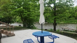 A blue picnic table with a white umbrella, surrounded by greenery and a river. - RV Bunkhouse on the River (Mason City)