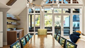 A dining area with a long wooden table, chairs with patterned cushions, and a view of a forest through large windows. - Arrabelle Premier Penthouse - ski in/ski out (Lionshead)