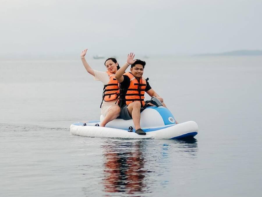 Two people on a watercraft with life jackets, floating on calm water.