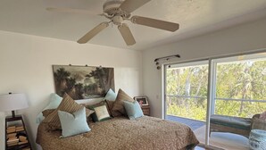 Une chambre avec un grand lit, un ventilateur de plafond, un tableau au mur et une vue sur l’extérieur grâce à une porte-fenêtre coulissante.