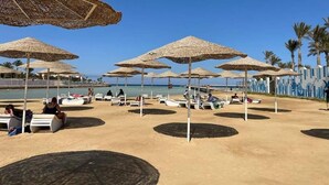 Een strand met ligbedden en rieten parasols, palmbomen en een helderblauwe lucht.