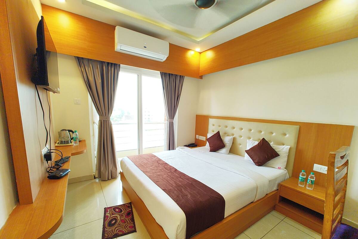 Quarto de hotel com uma cama grande, mesa de cabeceira com abajur, ar condicionado e janela com cortinas.