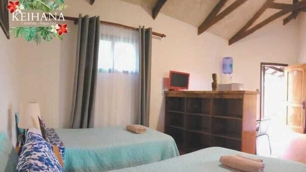 A room with two beds, a wooden cabinet, a television, and a window with curtains. - Keihana Rapanui (Hanga Roa)