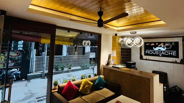 A cozy interior with a wooden ceiling, a sofa with colorful cushions, and a sign reading "MOUS JACHE". - Moustache Hostel Rishikesh (Rishikesh)
