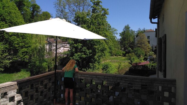 En person står på en stenbalkon med en hvid parasol og ser ud over et frodigt grønt landskab og en bygning med rødt tag.