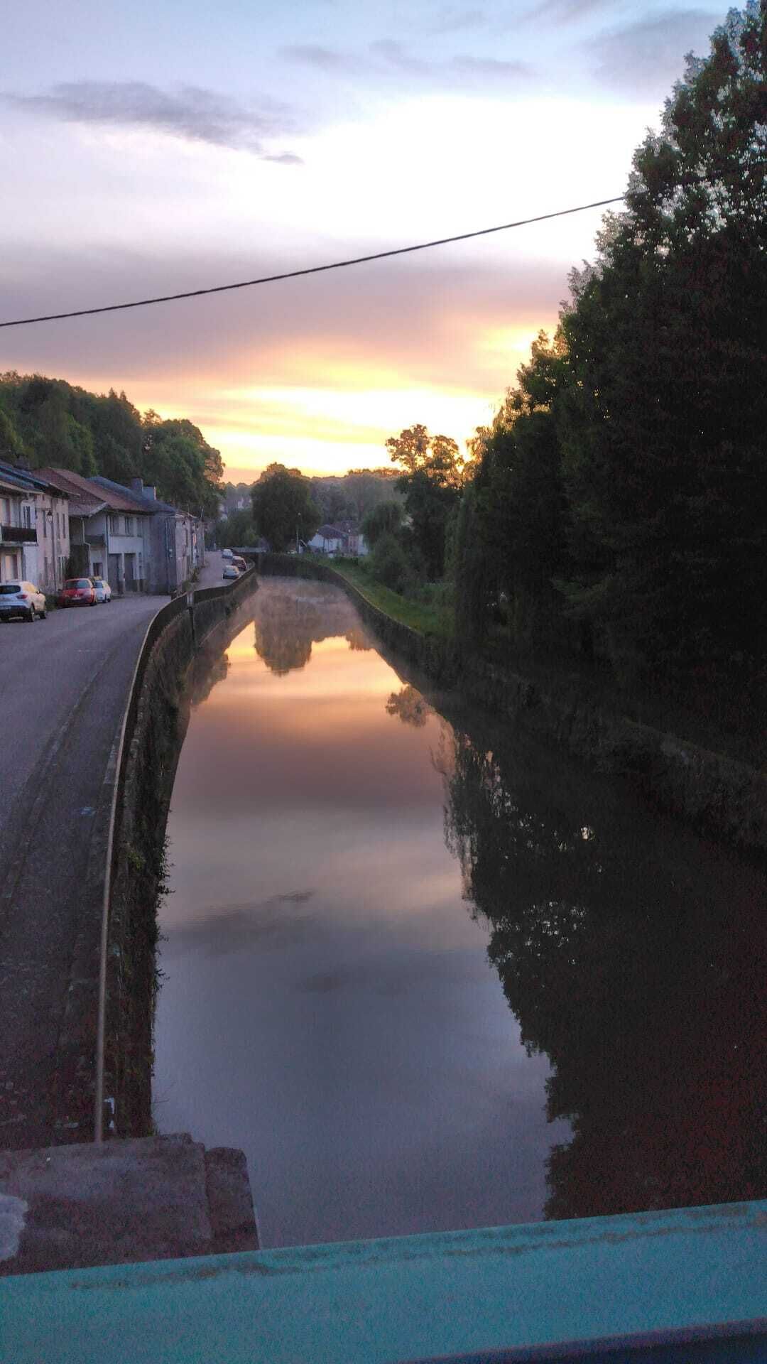 A canal with a calm water surface reflecting the sky, flanked by trees and a road with parked cars.