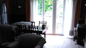 En stue med en mørk sofa, et træbord og et spisebord med stole. Et stort vindue med gardiner er synligt, og giver adgang til et udendørs område.