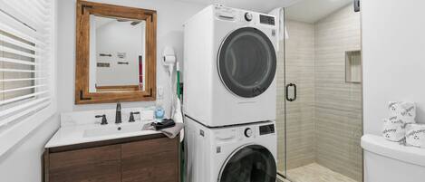 A compact bathroom with a washing machine, a sink, a mirror, and a shower area.