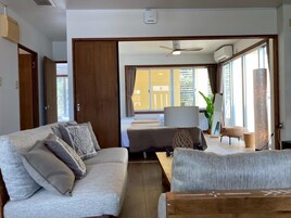 Egy modern nappali, amelyben kanapé, dohányasztal és étkezősarok található. Van egy nagy ablak, amelyen függöny van, egy mennyezeti ventilátor és egy légkondicionáló egység is.