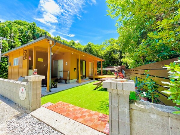 Un petit bâtiment en bois jaune, doté d’un porche abrité et d’une pelouse dallée.