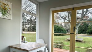 Una habitación con una mesa y silla blancos, un banco fuera y una puerta que da a un jardín.