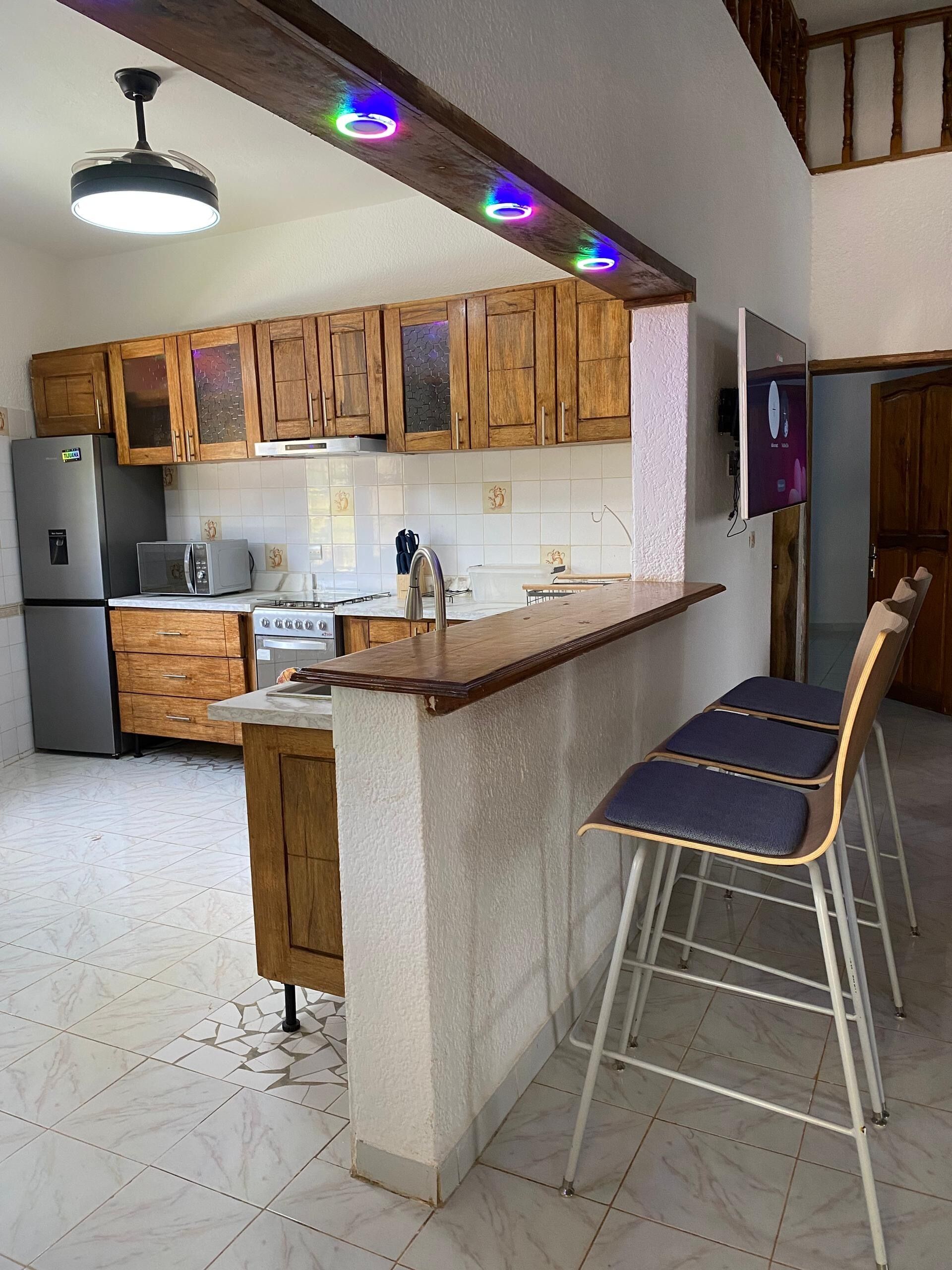 A kitchen with wooden cabinets, a built-in microwave, a refrigerator, a stove, and a bar area with stools.