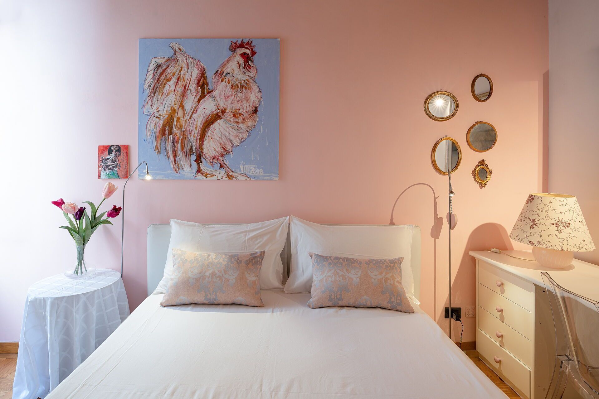 Une chambre à coucher avec un lit, des oreillers, une table de chevet avec une lampe, une commode, un miroir et un tableau accroché au mur.