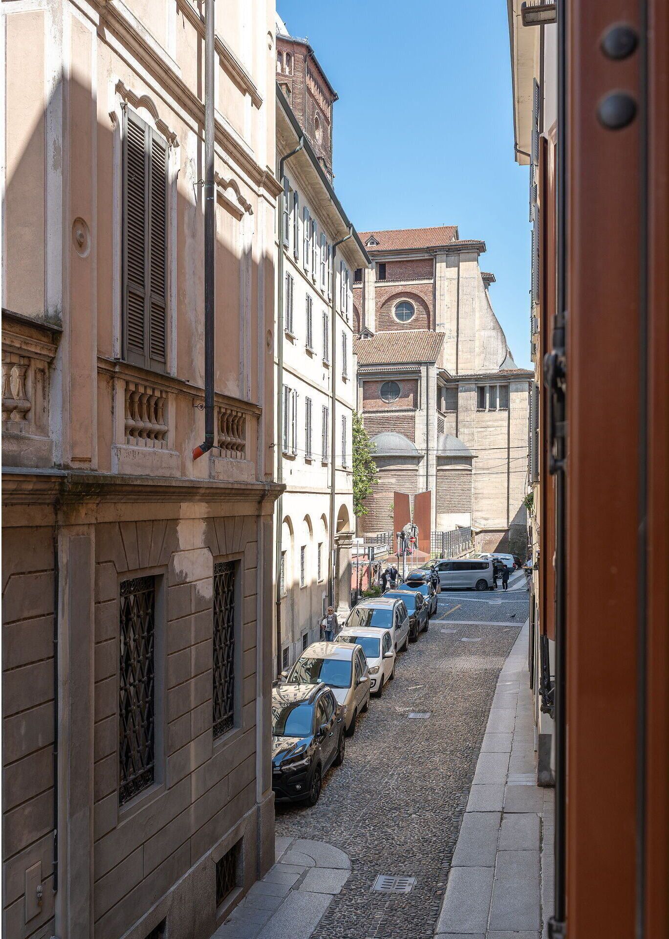 Une rue étroite bordée de bâtiments historiques, de voitures garées et d’un ciel d’un bleu éclatant.
