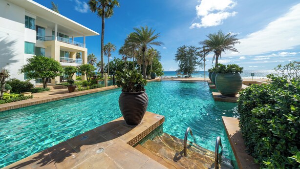 A clear blue pool with a palm tree and a large planter in the foreground, surrounded by a modern building with balconies and a view of the ocean. - Karon Beachfront Apartment 703 (Karon)