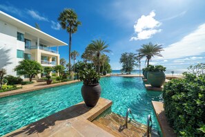 Piscina azul e límpida com uma palmeira e um grande vaso em primeiro plano, cercada por um edifício moderno com varandas e vista para o oceano.