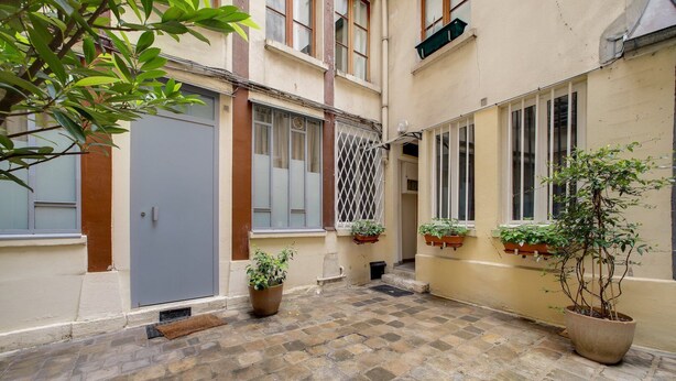A narrow courtyard with stone paving, a grey door, and potted plants.