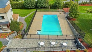 A rectangular swimming pool surrounded by a black metal fence, with a patio area featuring white round tables and chairs.