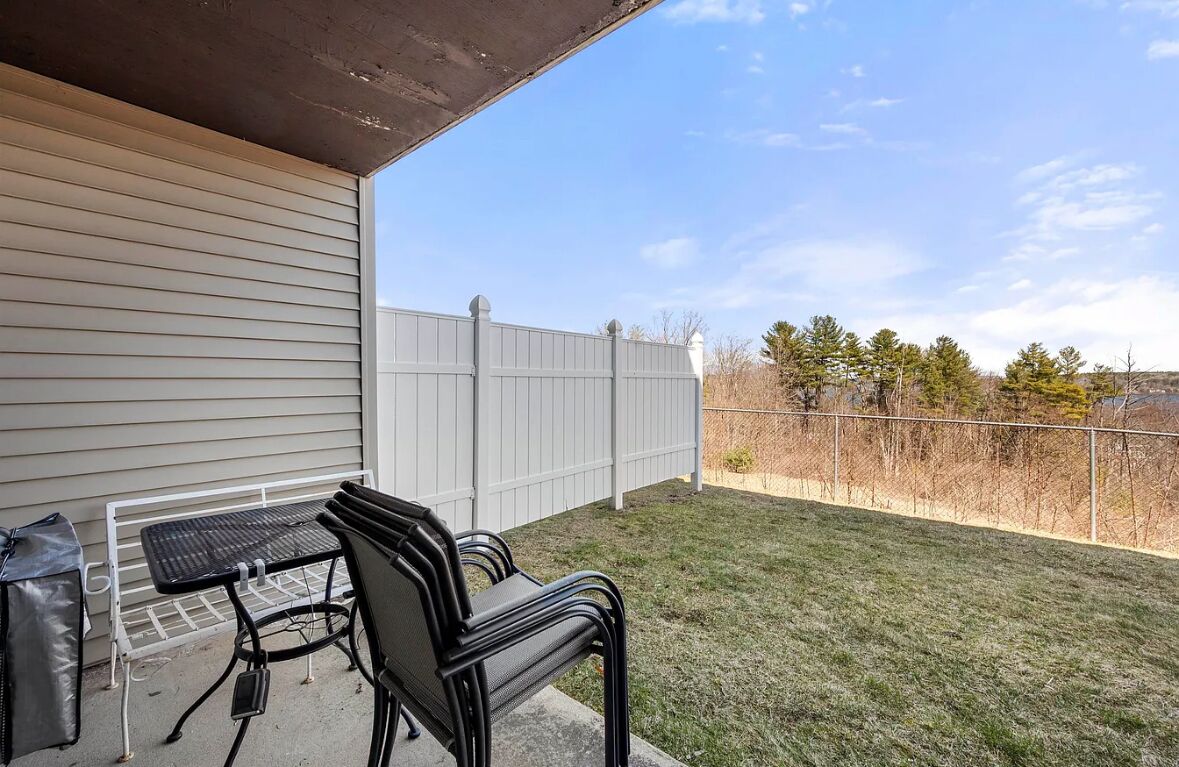 A patio with a table and chairs, a white fence, and a view of trees and hills.