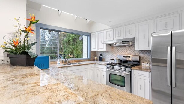 A modern kitchen with white cabinets, a stainless steel refrigerator, a gas stove, and a large window providing a view of the outdoors. - Luxury 5 Bedroom Villa w/Sauna (Vancouver)