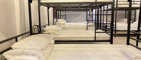 Una fila di letti a castello con biancheria e cuscini bianchi, ringhiere in metallo e un muro spoglio.