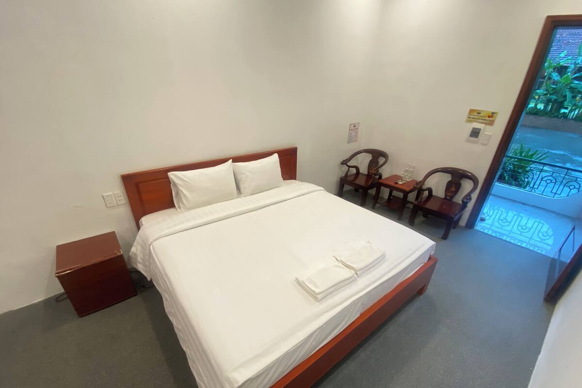 A hotel room with a single bed, a wooden nightstand, a small table with chairs, and a view of the outside balcony.
