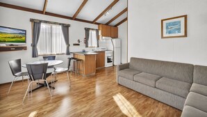 Een moderne keuken met houten balken, een zithoek met een ronde tafel en stoelen, een grijze bank en een schilderij aan de muur.