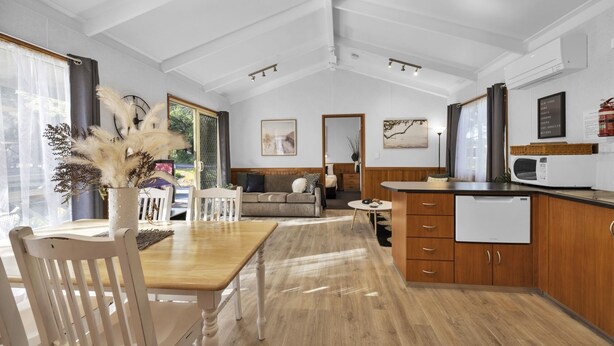 Een ruime kamer met een houten eettafel, witte stoelen, een bank en een keukenhoek met houten kasten en een magnetron.