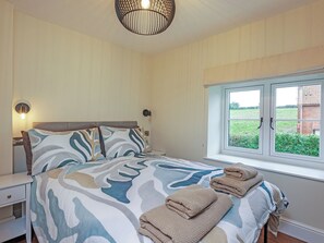 Ein Schlafzimmer mit Bett, Nachttischen, einem Fenster mit Blick auf Grünflächen und einer Deckenleuchte.